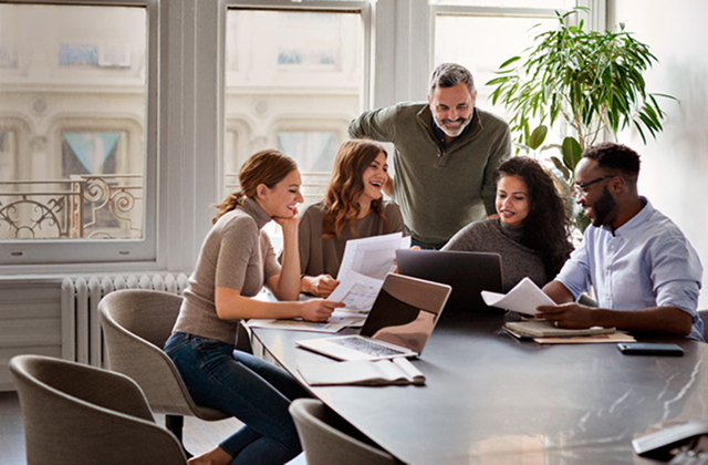 Group of colleagues collaborating around a table in a bright office, reviewing printed documents and using laptops during a team meeting